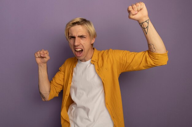 Excited young blonde guy wearing yellow t-shirt raising fists isolated on purple