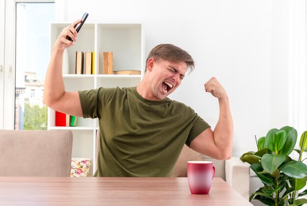 Excited young blonde handsome man sits at table with cup tenses biceps and holds phone inside living room