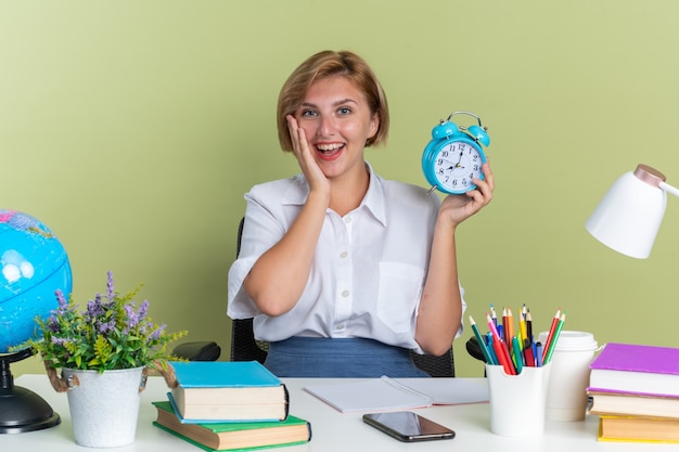 Excited young blonde student girl sitting at desk with school tools looking at camera keeping hand on face holding alarm clock isolated on olive green wall