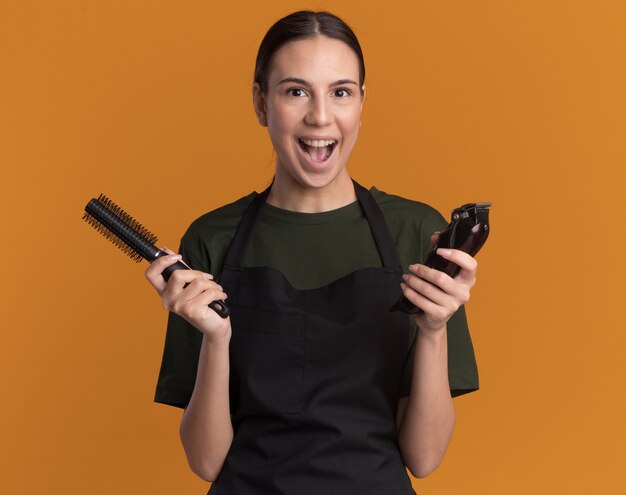 Excited young brunette barber girl in uniform holds hair clippers and comb isolated on orange wall with copy space