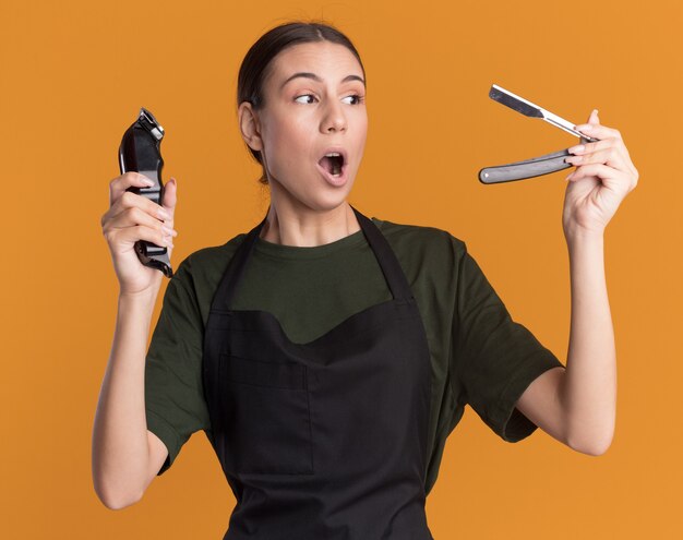 Excited young brunette barber girl in uniform holds hair clippers and looks at straight razor