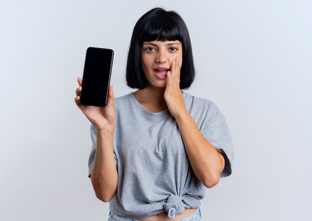 Excited young brunette caucasian woman puts hand on face and holds phone