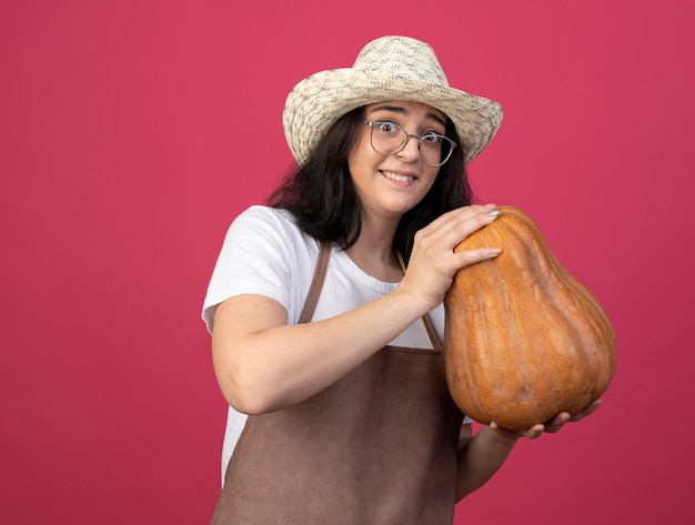 Excited young brunette female gardener in optical glasses and in uniform wearing gardening hat holds pumpkin isolated on pink wall with copy space