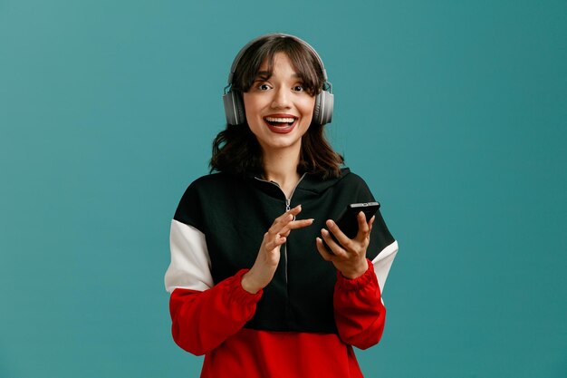 Excited young caucasian woman wearing headphones holding mobile phone looking at camera isolated on blue background