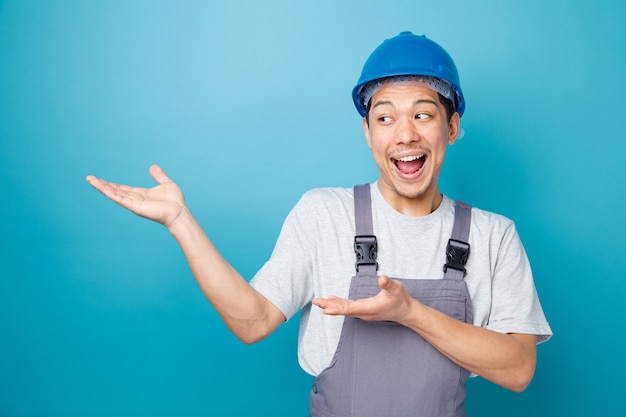 Excited young construction worker wearing safety helmet and uniform looking and pointing at side with hands 