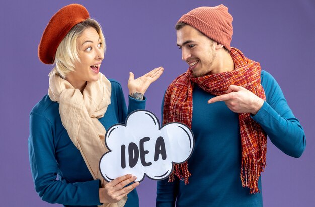 Excited young couple wearing hat with scarf on valentine's day holding and points at idea bubble isolated on blue background