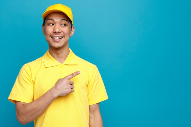 Excited young delivery man wearing uniform and cap looking and pointing at side 