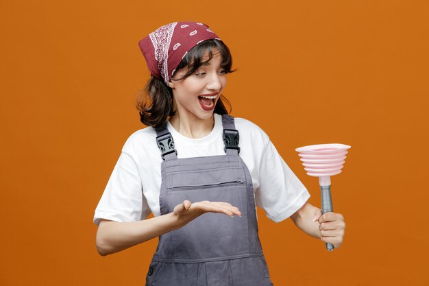 Excited young female cleaner wearing uniform and bandana holding plunger looking at it showing empty hand isolated on orange background