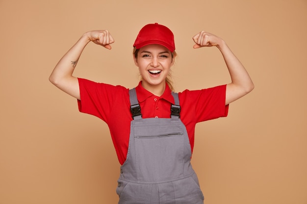 Free photo excited young female construction worker wearing uniform and cap doing strong gesture