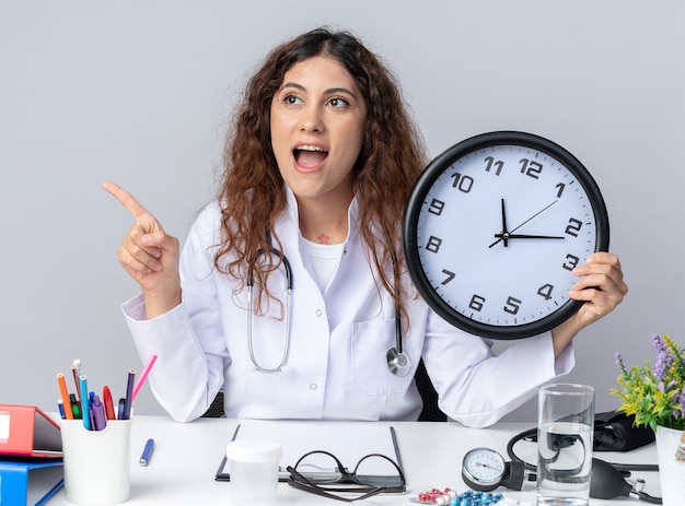 Excited young female doctor wearing medical robe and stethoscope sitting at table with medical tools holding clock looking and pointing at side isolated on white wall