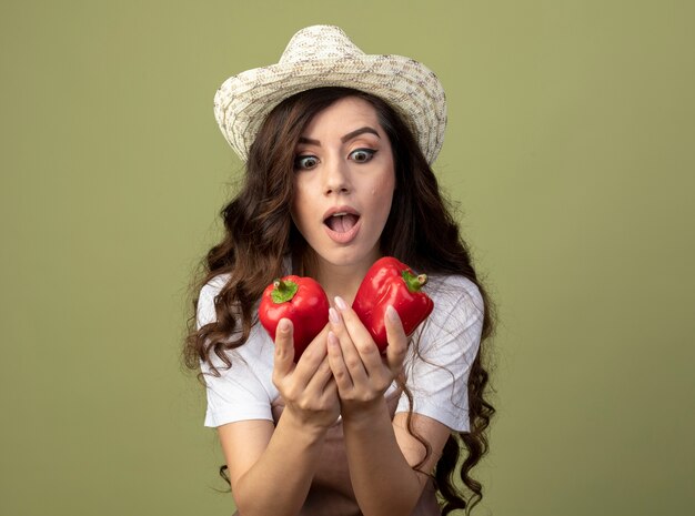 Excited young female gardener in uniform wearing gardening hat holds and looks at red peppers isolated on olive green wall