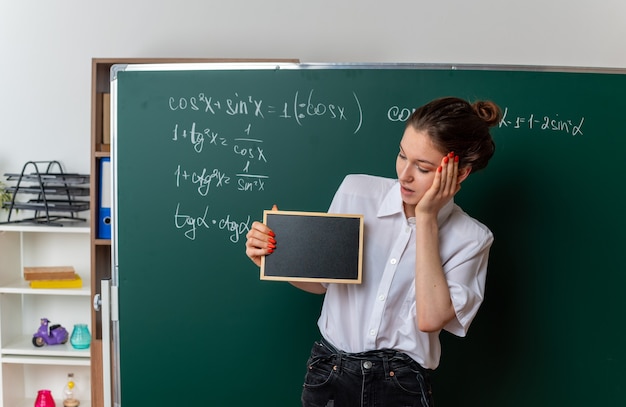 Free Photo excited young female math teacher standing in front of chalkboard showing and looking at mini blackboard keeping hand on face in classroom