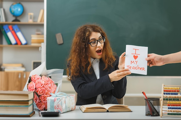 Free photo excited young female teacher received postcard sitting at desk with school tools in classroom