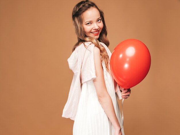 Excited young girl posing in trendy summer white dress. Woman model with red balloon posing. Ready for party