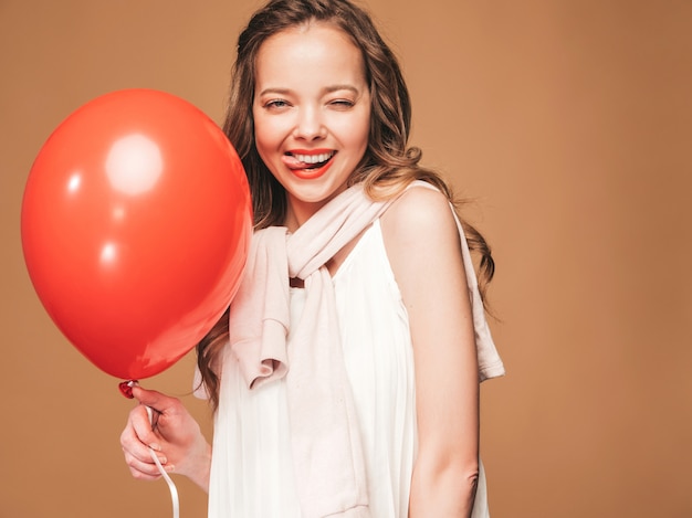 Excited young girl posing in trendy summer white dress. Woman model with red balloon posing.Showing her tongue and ready for party