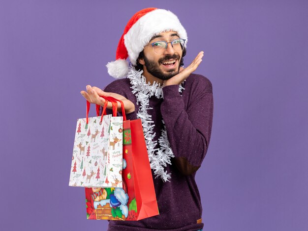 Excited young handsome guy wearing christmas hat with garland on neck holding gift bags spreading hands isolated on blue wall