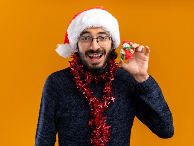 Excited young handsome guy wearing christmas hat with garland on neck holding toy isolated on orange wall