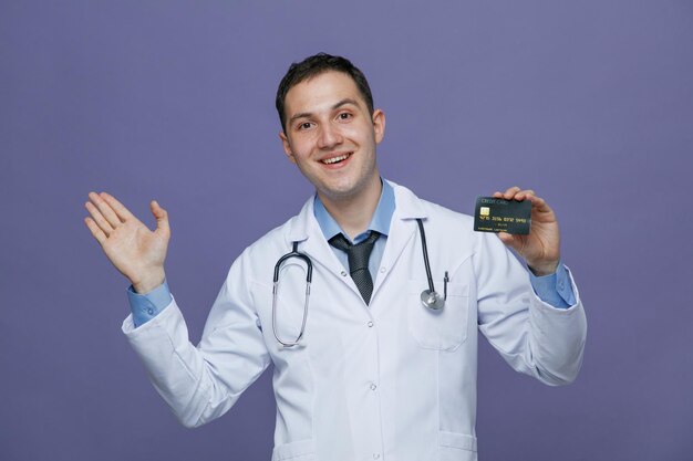 Excited young male doctor wearing medical robe and stethoscope around neck looking at camera showing credit card and empty hand isolated on purple background