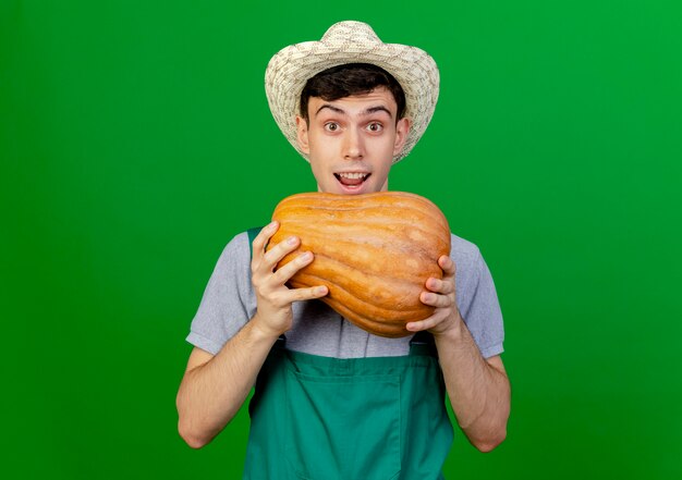 Excited young male gardener wearing gardening hat holds pumpkin 