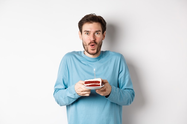 Excited young man making wish on birthday cake with lit candle, celebrating bday, standing on white background.