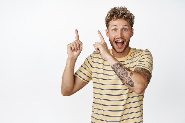 Excited young modern man with curly blond hairstyle pointing fingers at upper left corner smiling and staring amazed showing happy news white background