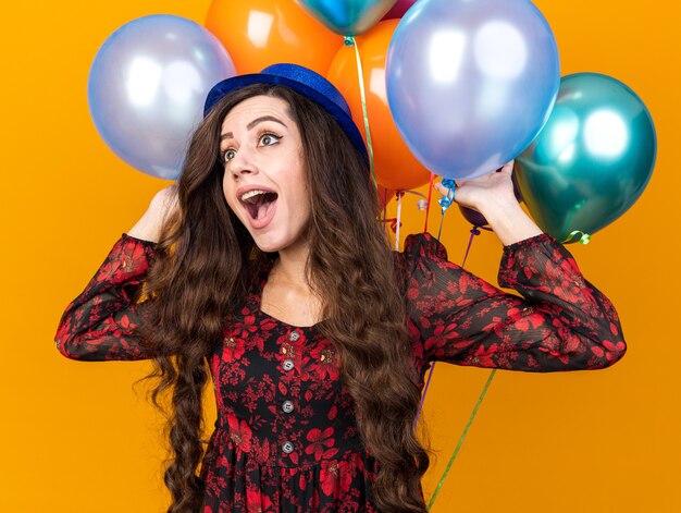 Excited young party girl wearing party hat standing in front of balloons touching them looking at side isolated on orange wall