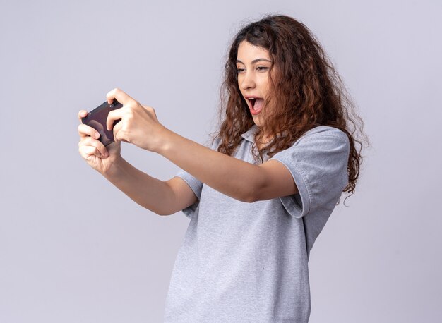 Excited young pretty caucasian girl standing in profile view holding mobile phone playing game on phone isolated on white wall