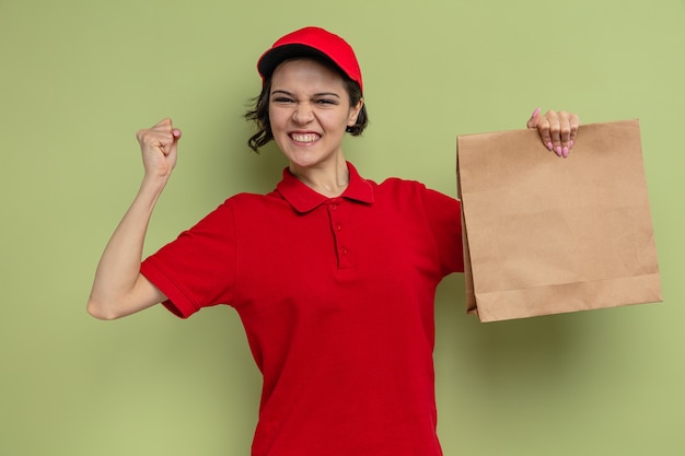 Excited young pretty delivery woman holding paper food packaging and raising fist up 