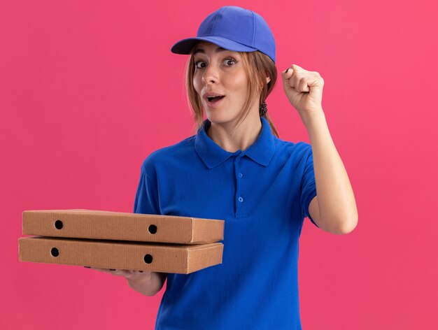 Excited young pretty delivery woman in uniform raises fist up and holds pizza boxes isolated on pink wall