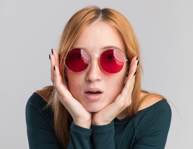 Excited young redhead ginger girl with freckles in sun glasses puts hands on chin  isolated on white wall with copy space