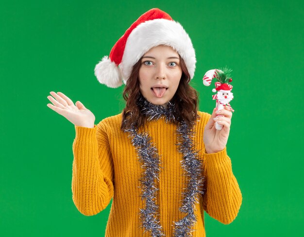 Excited young slavic girl with santa hat and with garland around neck holding candy cane and keeping hand open 