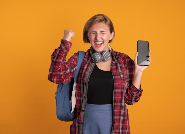 Excited young slavic student girl with headphones wearing backpack stands with closed eyes raising fist up holding phone 