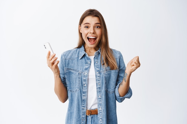 Excited young woman scream yes and smiling amazed, holding smartphone and staring at front, winning online, standing happy with achievement against white wall