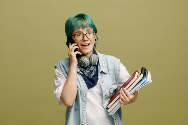 Excited young woman wearing glasses headphones and bandana on neck holding note book and note pads looking at side while talking on phone isolated on olive green background