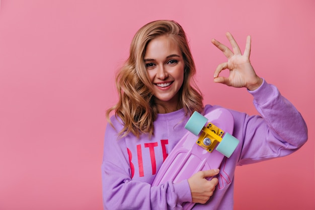 Excited young woman with blonde wavy hair posing with skateboard. Relaxed smiling girl in purple shirt showing okay sign.