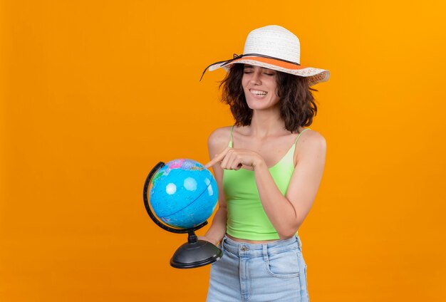 An excited young woman with short hair in green crop top wearing sun hat pointing at a globe with index finger 