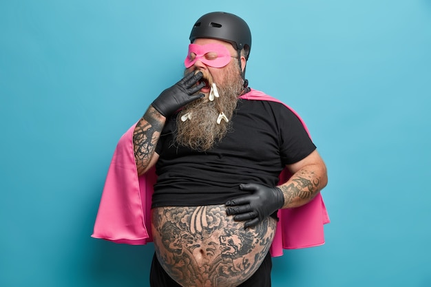 Free photo exhausted male superhero yawns and covers mouth stands with big tattooed belly indoor being tired of doing different things models against blue wall