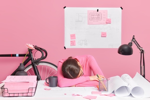 Free Photo exhausted tired overworked female engineer works all day on new design project leans at table wants to sleep surrounded with sketches and blueprints poses in home office. lack of productivity