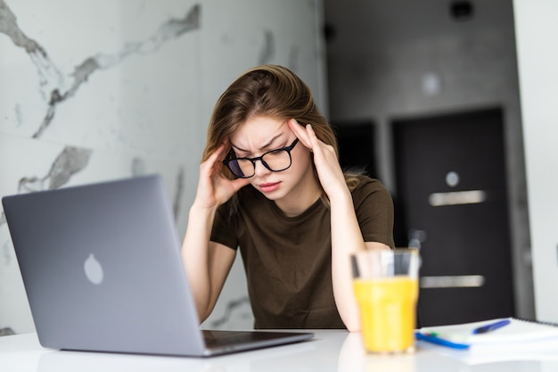 Exhausted tired woman working using on laptop holding head in hand sitting in kitchen at home