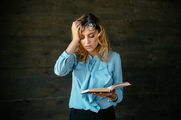 Exhausted woman looks tired, holds a book, keeps her head with hand