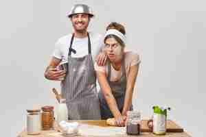 Free photo exhausted woman with flour on face, kneads dough, tired of preparing homemade bread, cheerful man in apron leans on shoulder, holds bowl, glad to help wife on kitchen. couple make pizza together