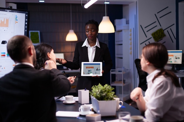 Exhausted workaholic african american showing financial graphs using tablet overworking in company o...