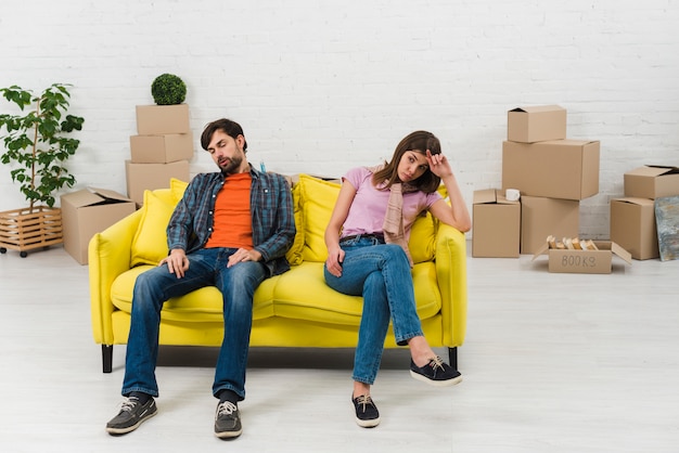 Exhausted young couple sitting on yellow sofa with moving cardboard boxes in their new home