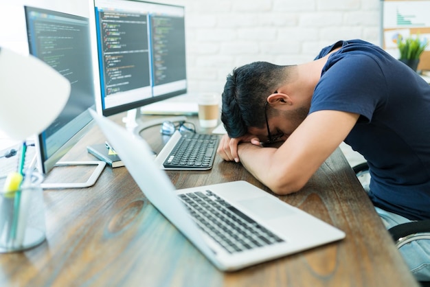 Exhausted young male software developer sleeping by technology while leaning on desk