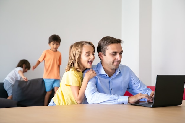 Exited lovely girl looking at laptop screen and hugging dad. Caucasian middle-aged father working at home when cute children playing on sofa. Childhood, fatherhood and digital technology concept