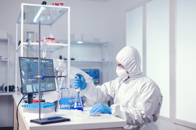 Experienced biologist dressed in ppe holding pipette with blue solution. Chemist in modern laboratory doing research using dispenser during global epidemic with covid-19.