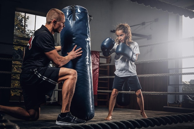 Experienced boxer trainer is training new little girl boxer for special competitions using punching bag.