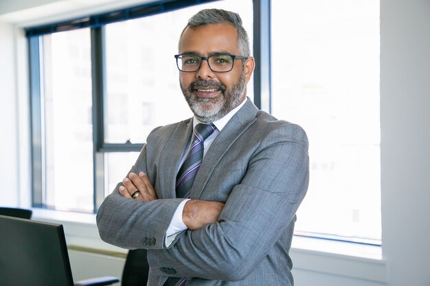 Experienced businessman standing in office room. Indian content office employee in eyeglasses smiling and posing with folded hands. Business, management and corporation concept