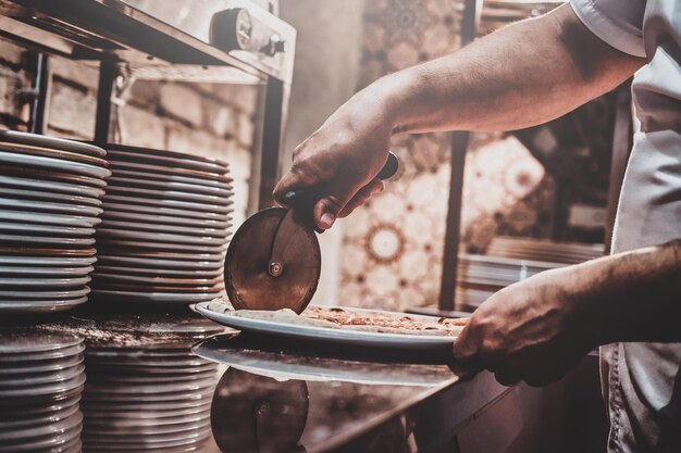 Experienced chef is cutting freshly prepared pizza with special knife.