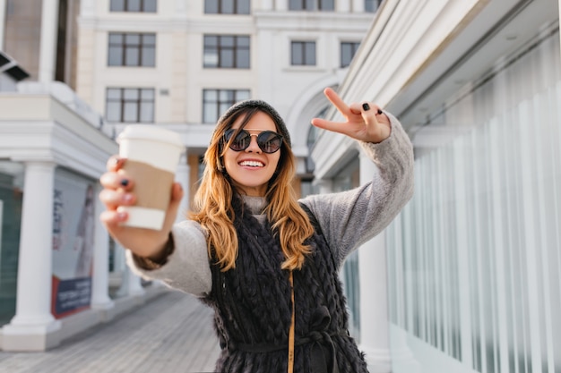 Expressing brightful positive emotions of fashionable city woman stretching coffee to go on sunny street. Beautiful smiling woman in modern sunglasses, knitted hat having fun outdoor.